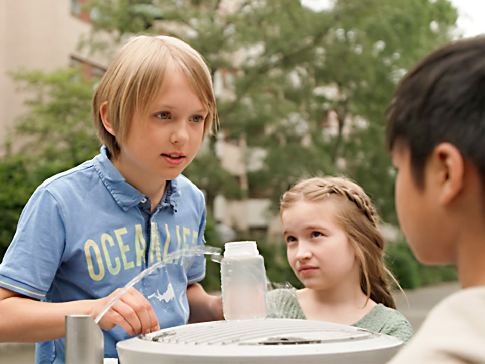 Kinder stehen an einem Trinkbrunnen und unterhalten sich über Wasser